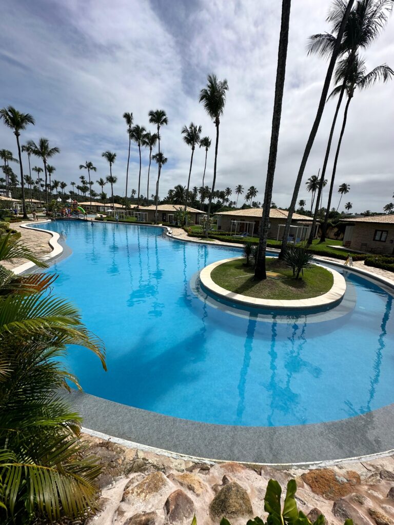 Uma piscina coletiva com árvores e pequenas casas ao redor localizada em um Resort no Nordeste - reforma-de-piscina-coletiva-em-resort